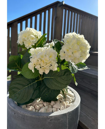 De Kunstplant Hortensia Wit UV 35 cm, met witte bloemen en groene bladeren, staat op een zonnige dag buiten op witte kiezelstenen, met op de achtergrond houten terrasleuningen en een trap.