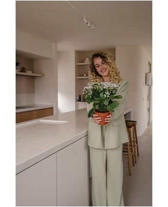 Een vrouw met krullend blond haar in een lichtgroen pak glimlacht terwijl ze de Kunstbloem Kalanchoë plant UV 31 cm wit vasthoudt in een moderne, neutraal getinte keuken met open planken.