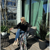 Een vrouw met zonnebril, zwart topje en blauwe jeans ontspant op een kussensstoel op een zonnig balkon met een drankje in de hand. De ruimte is voorzien van de Kunstplant Laurier UV d33 cm, een levendig decor, een kleurrijke deken en een kleine tafel met planten.