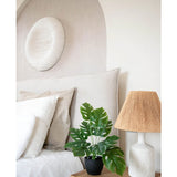 Een moderne slaapkamer in neutrale tinten heeft een wit bed met beige en witte kussens, een houten nachtkastje met een lamp van keramiek en een geweven kap, abstracte muurkunst boven het bed en de Kunst Monstera 40 cm in sierpot zwart die groen toevoegt.