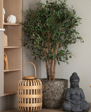 De Kunst olijfboom 120 cm UV staat, samen met een houten lantaarn en een Boeddha, op de vloer naast een lichte houten plank vol decoratie. De sfeer is sereen en minimalistisch dankzij deze onderhoudsarme blikvanger.