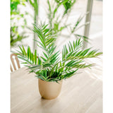 Een Kunstpalm Parlour UV 60 cm ligt op een lichte houten tafel, zonlicht stroomt naar binnen door de grote ramen terwijl andere kunstplanten zacht vaag op de achtergrond verschijnen.
