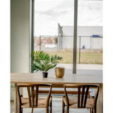Een houten tafel met twee stoelen, een Kunstpalm Parlour UV 60 cm in een pot en een gouden vaas staan bij een groot raam met uitzicht op een industriegebied met gebouwen en een hek.