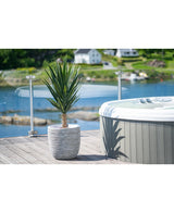 Een Kunstplant Yucca 100 cm UV staat op een houten dek naast een hot tub met glazen reling, met uitzicht op een rustig meer en groene heuvels bezaaid met huizen.