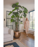 Een vrouw in een beige outfit staat binnen naast de Kunstplant Monstera 240 cm in een bronzen pot. De moderne woonkamer heeft een witte bank, grote ramen met luxaflex en een houten vloer.