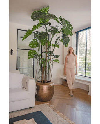 Een vrouw in een beige outfit staat binnen naast de Kunstplant Monstera 240 cm in een bronzen pot. De moderne woonkamer heeft een witte bank, grote ramen met luxaflex en een houten vloer.