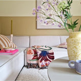 Een gezellige woonkamer met een lichtbeige sofa, een kleurrijke deken met bloemen en kussens met een patroon. Op een ronde tafel staan een gele vaas met Kunsttak Daphne Spray 108 cm paars en een klein blauw kopje. Aan de neutraal getinte muur hangt kunst.