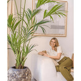 Een vrouw met blond haar, in een crèmekleurige top en olijfgroene broek, zit glimlachend op een witte bank en houdt een mok vast. De Kunstpalm Kentia 220 cm versterkt de tropische sfeer van de lichte, gezellige kamer met abstracte wandkunst.