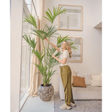 In een lichte woonkamer staat een vrouw naast de Kunstpalm Kentia 220 cm, terwijl ze zachtjes de bladeren aanraakt. Het tropische interieur heeft een lichte bank met kussens, ingelijste muurkunst en grote ramen die natuurlijk licht binnenlaten.