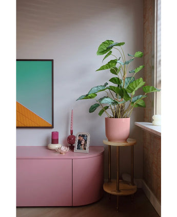 Een moderne woonkamerhoek met een roze dressoir, kaarsen, geometrische muurkunst, een ingelijste foto en de Kunstplant Appelblad 90 cm-een grote bladplant in een roze pot op een kleine houten standaard bij het raam.