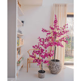Twee Kunstplant Bougainvillea 125 cm Paars met levendige paarse bloemen in potten met structuur fleuren een moderne kamer op, naast een houten stoel, boekenplank en een groot raam met beige gordijnen.