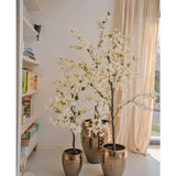 Een Kunstplant Bougainvillea van 150 cm groot staat in een decoratieve gouden pot bij een zonovergoten raam met beige gordijnen en voegt elegantie toe aan de gezellige ruimte. Nabijgelegen boekenplanken versterken de uitnodigende interieursfeer.