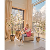 Een vrouw in een roze vest met patroon en spijkerbroek zit op een beige fauteuil bij grote ramen met crèmekleurige gordijnen, omringd door drie Kunstplant Bougainvillea van 180 cm wit in glanzend gouden plantenbakken. Zonlicht vult de moderne, luchtige kamer.