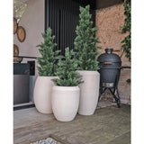 Drie hoge, witte plantenbakken met Kunstplant Conifeer UV 90cm staan op een houten terras naast een modern gebouw. Een zwart keramisch rooster staat vlakbij tegen een stenen muur met wijnranken.