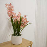 De Kunstplant Cymbidium 58 cm roze, met hoge groene bladeren en roze orchideeachtige bloemen, staat op een lichte houten tafel tegen een effen witte muur en een wit gordijn.