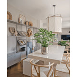 Een vrouw glimlacht in een lichte, moderne eetkamer met houten stoelen, een witte tafel met de Kunstplant Dieffenbachia 75 cm als middelpunt en open planken versierd met boeken en kunst.