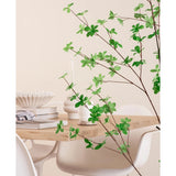 Een moderne eethoek met witte stoelen en een lichthouten tafel, gedecoreerd met boeken, kaarsen en een witte vaas, wordt op de voorgrond omlijst door de Kunstplant Enkianthus 150 cm bladgroene plant.