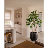 Een moderne, minimalistische keuken en eethoek in neutrale tinten heeft open planken, een gebogen eiland met een houten kruk, een houten eetset en de Kunstplant Ficus 210 cm als stijlvolle potplant. Natuurlijk licht verlicht de ruimte.