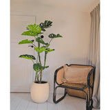 Een Kunstplant Monstera 150 cm staat naast een rotan stoel met een bruin kussen in een lichte, minimalistische kamer met witte muren, lichthouten vloeren en beige gordijnen.