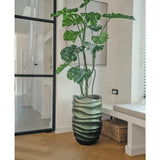 De Kunstplant Monstera 150 cm, met zijn grote groene bladeren, staat in een pot met golvend patroon op een houten vloer in een moderne, lichte kamer met een glazen deur, ingelijste kunst en een rieten mand in de buurt.