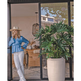 Een vrouw in een blauwe blouse, witte broek en hoed lacht bij een glazen deur naast de Kunstplant Philodendron 125 cm in een beige pot. Rieten stoelen en decor zijn zichtbaar in de kamer.