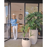 Een vrouw met hoed, blauwe blouse en witte broek leunt tegen een glazen deur. Op de voorgrond staat de Kunstplant Philodendron 125 cm. Binnenin is een gezellige, moderne leefruimte te zien.