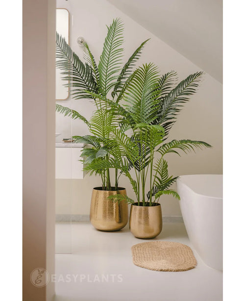 Een Kunstplant Phoenix 180 cm met weelderige groene bladeren in een glanzende gouden pot staat naast een wit bad en een beige geweven mat in een moderne, minimalistische badkamer, aangevuld met een spiegel en een raam voor een serene uitstraling.