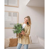 Een vrouw in een beige blazer en spijkerbroek staat in een moderne woonkamer en glimlacht terwijl ze een Kunstplant Podocarpus 47 cm vasthoudt. Abstracte kunst siert de lichte muur boven een crèmekleurige bank met bruine kussens.