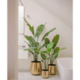 Een Kunstplant Strelitzia Nicolai Deluxe 120 cm staat in een glanzende gouden plantenbak op de vloer van een moderne, minimalistische badkamer met een wit bad en lichtgekleurde muren.