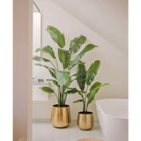 Twee Kunstplant Strelitzia Nicolai Deluxe 180 cm in glanzend gouden potten staan bij een wit bad in een moderne, minimalistische badkamer met een klein rond geweven vloerkleed op de vloer.