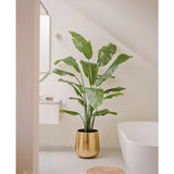 De Kunstplant Strelitzia Nicolai Deluxe 180 cm in een glanzende gouden pot staat op een witte vloer naast een geweven vloerkleed in een lichte, moderne badkamer met witte muren, een bad en een wastafel aan de muur.