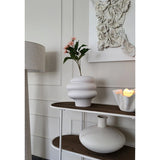 Een modern interieur met een wit wandkunstwerk met textuur, een witte sculpturale vaas met Kunsttak Bloeiende Eucalyptus 62 cm oud roze, een decoratieve kaars en een ronde keramische vaas op een houten plank naast een staande lamp.
