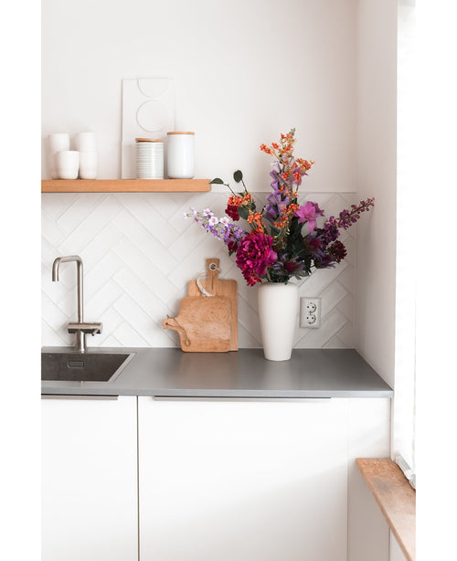 Een moderne witte keuken met een grijs aanrechtblad, een gootsteen, een houten snijplank en een witte vaas gevuld met Kunstbloem Hortensia 55 cm donkerroze. Witte kopjes en potten staan op een houten plank boven het aanrecht.