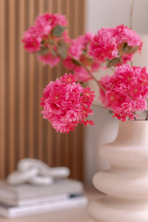 Een witte vaas van keramiek houdt een Kunsttak Kersenbloesem 85 cm roze vast. De achtergrond toont verticale houten panelen, een stapel boeken en een klein decoratief object bovenop.