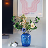 Een blauwe glazen vaas met een Kunsttak Berry 76 cm paars kunstbloem staat op een witte kast. Ernaast staan twee witte pompoenen met gouden stelen. Aan de muur erachter hangen een tv en een abstracte roze kunstprint.