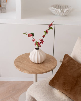 Op een klein rond tafeltje staat de Kunsttak Berries 64 cm roze – een witte vaas gevuld met roze kunstbessentakjes en groene blaadjes – naast een beige stoel met een bruin kussen in een gezellige, minimalistische kamer.