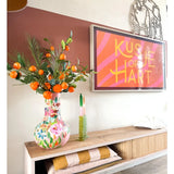 Op een houten plank staat een kleurrijke vaas met daarin de Kunsttak Mandarijn 93 cm oranje, naast een ingelijst kunstwerk met roze strepen waarop „Kusje op je Hart” staat. Op de plank liggen ook een groene kaars en opgevouwen dekens.