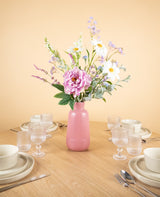 Op een ronde houten tafel met vier couverts staat het Zijden kunstbloemen boeket Misty Minthe in een roze vaas in het midden, waardoor het tafereel oplicht tegen een lichtbeige achtergrond.