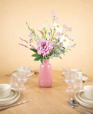 Op een ronde houten tafel met vier couverts staat het Zijden kunstbloemen boeket Misty Minthe in een roze vaas in het midden, waardoor het tafereel oplicht tegen een lichtbeige achtergrond.