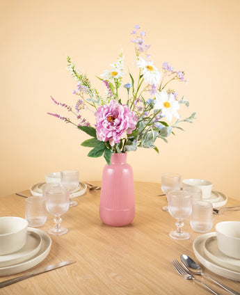 Op een ronde houten tafel met vier couverts staat het Zijden kunstbloemen boeket Misty Minthe in een roze vaas in het midden, waardoor het tafereel oplicht tegen een lichtbeige achtergrond.