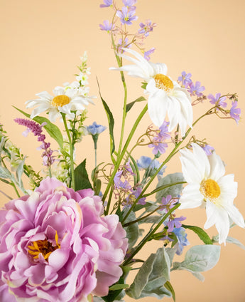 Het Zijden kunstbloemen boeket Misty Minthe heeft witte margrieten, paarse en blauwe bloesems en een grote roze pioenroos tegen een zacht perzikkleurige achtergrond.