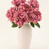 De Zijden kunstbloemen boeket Peony Dusty Pink is gevat in een witte vaas van keramiek met textuur. De vaas heeft grote roze pioenrozen met weelderige groene bladeren tegen een lichte, effen achtergrond.
