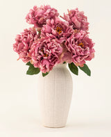 De Zijden kunstbloemen boeket Peony Dusty Pink is gevat in een witte vaas van keramiek met textuur. De vaas heeft grote roze pioenrozen met weelderige groene bladeren tegen een lichte, effen achtergrond.