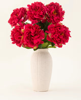 Een vaas van wit keramiek met textuur bevat zes grote, levendige zijden bloemen uit het Zijden kunstbloemen boeket Peony Raspberry Red, met weelderige groene bladeren, tegen een effen lichte achtergrond.