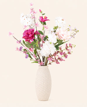 Een vaas met beige structuur bevat het Zijden kunstbloemen boeket Pink Reverie - een arrangement van roze, paarse en witte kunstbloemen met groene bladeren tegen een effen lichte achtergrond.