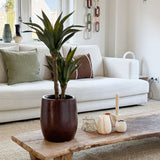 Een moderne woonkamer met een witte bank, groene en bruine kussens, een houten salontafel versierd met kaarsen en de Kunstplant Agave 76 cm, en een wanddecoratie van kettinggaas. Door de ramen achter de bank valt natuurlijk licht naar binnen.
