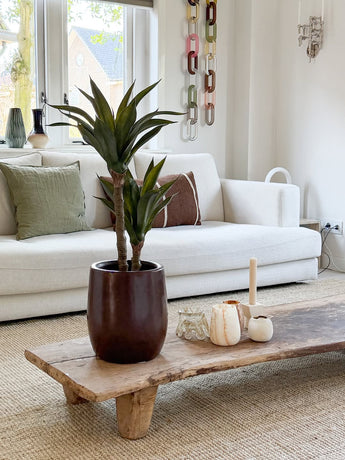 Een moderne woonkamer met een witte bank, groene en bruine kussens, een houten salontafel versierd met kaarsen en de Kunstplant Agave 76 cm, en een wanddecoratie van kettinggaas. Door de ramen achter de bank valt natuurlijk licht naar binnen.