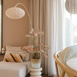 Een gezellige zithoek met een beige chaise longue, een gestreept kussen, de Kunstplant Caladium 90 cm roze die voor een vleugje groen zorgt, een moderne lamp, lichte gordijnen en een houten vloer. Zacht natuurlijk licht valt door de transparante gordijnen naar binnen.