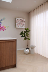 Een moderne, minimale hoek met de Kunstplant Ficus Tabaksplant Deluxe 155 cm, een klein wit krukje, ronde zilveren plantenbak, beige gordijnen, houten kast, muurkunst en vaas met bloemen op een lichthouten vloer.