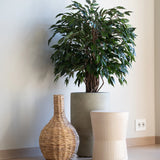 Een Kunstplant Ficus Liana 90 cm staat in een grijze plantenbak naast een gevlochten vaas en een licht geribbeld krukje, alles op een houten vloer tegen een effen witte muur.